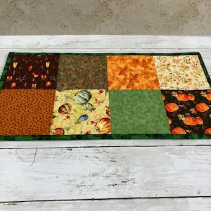 May include: A rectangular quilted table runner featuring eight distinct fabric panels with autumn-themed patterns, including pumpkins, leaves, and foliage in shades of orange, brown, green, and cream, all bordered by a dark green fabric trim.