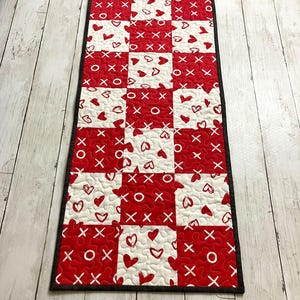 May include: A rectangular, quilted table runner with a repeating pattern of red and white squares. Each square features hearts, X's, and O's. The runner is bordered with a thin black trim and is placed on a light wood surface.