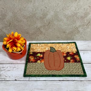 May include: A rectangular fabric placemat featuring a central orange gingham pumpkin appliqué on a dark red background with small pumpkins, bordered by tan pumpkin-print fabric and a geometric tan pattern, finished with a dark green fabric trim.