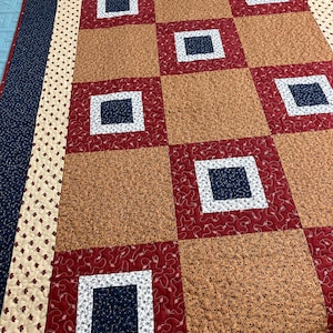 May include: A patchwork quilt featuring a repeating grid of squares in shades of deep red, tan, and navy blue. The design includes smaller navy blue squares centered within red blocks, surrounded by a cream border with a small red floral pattern.