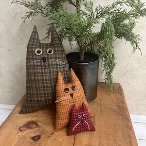 May include: Three handmade cat-shaped fabric pillows in varying sizes. The largest is plaid in brown and navy blue, the medium is orange plaid, and the smallest is red plaid. Each has button eyes and stitched details.