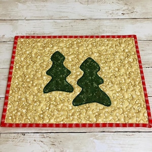 May include: A quilted placemat featuring two green Christmas tree appliqués on a gold background with white snowflake patterns. The placemat has a red and white striped border, perfect for holiday decor.