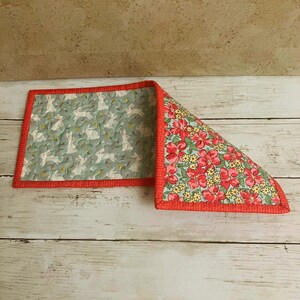 May include: A quilted placemat with a red border. One side features a pattern of white rabbits on a light green background. The other side displays a floral pattern with red flowers and yellow accents on a light green background.
