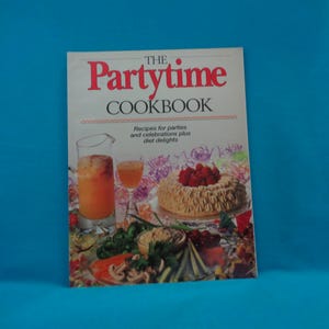 May include: The Partytime Cookbook, featuring recipes for parties and celebrations, is displayed against a blue background. The cover has the title in red letters. The image includes a cake, drinks, and other party food.