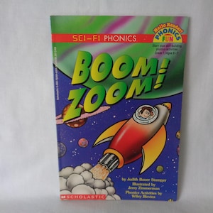 May include: A colorful book cover with the title "BOOM! ZOOM!" in green and yellow letters. The book is part of the "Sci-Fi Phonics" series and features a cartoon rocket ship with a young boy inside. The book is published by Scholastic.