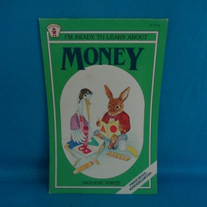May include: A green book cover with the title "I'm Ready to Learn About Money" in white text. The cover features an illustration of a stork and a rabbit holding coins. The book is for preschool and kindergarten.