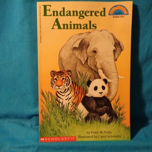May include: A book cover with a yellow background and the title "Endangered Animals" in large, bold, black letters. The cover features illustrations of an elephant, a tiger, and a panda bear in a grassy setting. The book is part of the "Hello Reader" series for grades 1 and 2.