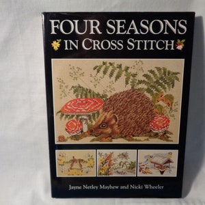 May include: Book cover with the title "Four Seasons in Cross Stitch" and a colorful cross stitch design of a hedgehog in a forest setting. The book is by Jayne Netley Mayhew and Nicki Wheeler.