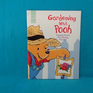 May include: A softcover book titled Gardening With Pooh, Cheerful Poems Plus Planting Pointers, featuring an illustration of Winnie the Pooh wearing a straw hat and blue overalls, holding a small sign decorated with colourful flowers.