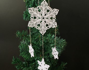 May include: White lace snowflake ornament hanging from a green Christmas tree. The snowflake has a delicate design and is attached to three smaller snowflakes.