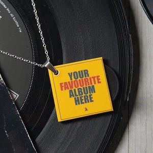 May include: A silver-toned metal chain necklace featuring a square pendant with a yellow background and the text "YOUR FAVOURITE ALBUM HERE" printed in red and dark grey. The pendant is positioned over a black vinyl record with visible grooves.