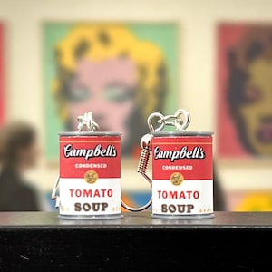 May include: A pair of silver earrings shaped like Campbell's Soup cans. The cans are red with white lettering that reads "Campbell's Condensed Tomato Soup".