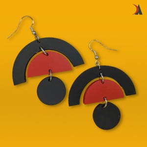 May include: A pair of contemporary dangle earrings. Each earring has a black arch, a red semi-circle, and a black circle. The earrings are suspended from silver-tone hooks, set against a yellow backdrop.