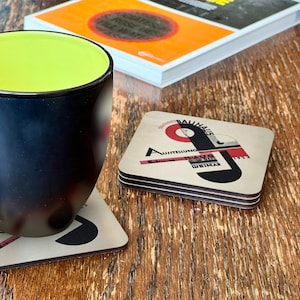 May include: A set of four square coasters featuring a Bauhaus design with the text "Austellung D-A-U-O 19-11-1923 Weimar" in black and red lettering on a white background.