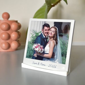 May include: A white framed photo print of a wedding couple, with the text "Sam & Fiona - 2026" at the bottom. The photo is displayed in a white stand. A terracotta-coloured decorative object is in the background.