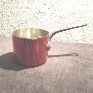 May include: A small, red saucepan with a silver interior and a long, dark handle. The handle extends horizontally from the side of the pan. The pan is sitting on a gray surface.