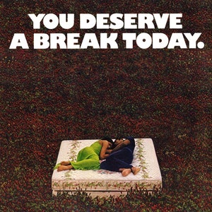 May include: A couple are lying on a white mattress with a floral print, surrounded by a field of red clover. The text "YOU DESERVE A BREAK TODAY." is displayed above the mattress.