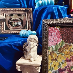 Pu&ograve; includere: Una natura morta con uno sfondo in tessuto di raso blu, un ritratto incorniciato di un uomo sorridente con denti d'oro, una statua di cherubino bianca e una stampa floreale incorniciata con fiori rosa, gialli e rossi.