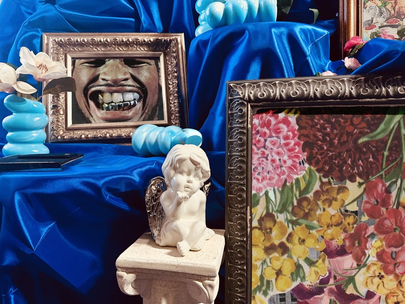 Pu&ograve; includere: Una natura morta con uno sfondo in tessuto di raso blu, un ritratto incorniciato di un uomo sorridente con denti d'oro, una statua di cherubino bianca e una stampa floreale incorniciata con fiori rosa, gialli e rossi.