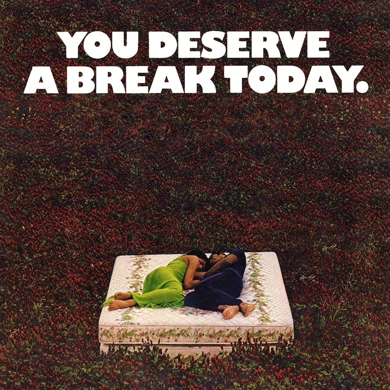 May include: A couple are lying on a white mattress with a floral print, surrounded by a field of red clover. The text "YOU DESERVE A BREAK TODAY." is displayed above the mattress.