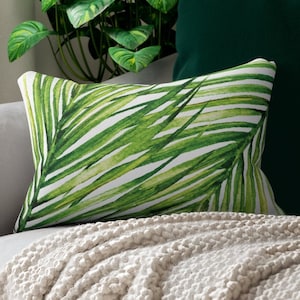 May include: A rectangular cushion featuring a green palm leaf design, set against a grey sofa with a cream knitted throw. A dark green cushion and a potted plant are in the background, creating a natural theme.