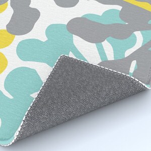 May include: A rug with a repeating pattern of abstract shapes in yellow, gray, and teal on a white background. The rug's edge is visible, showing a gray backing.