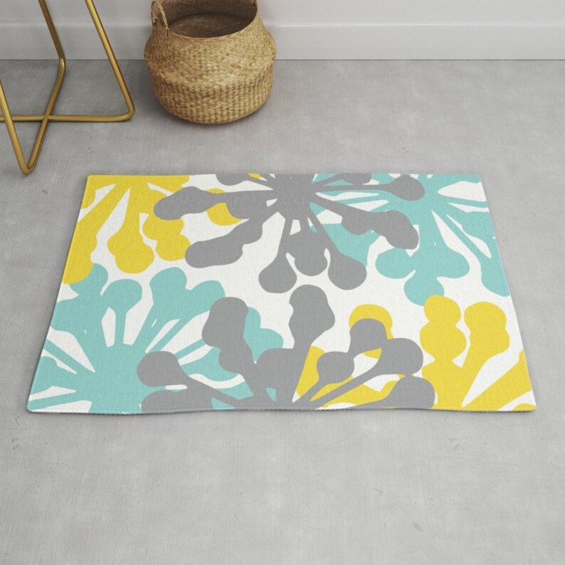 May include: A rectangular rug with a floral pattern in yellow, gray, and teal on a white background. The rug is on a light gray floor, with a gold-colored side table and a woven basket in the background.