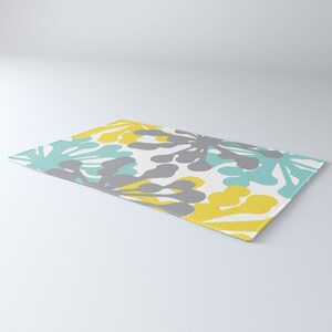 May include: A rectangular rug with a repeating floral pattern in shades of gray, yellow, and teal on a white background. The rug's design features abstract, stylized flower shapes, creating a modern and colorful aesthetic. The rug is suitable for indoor use.