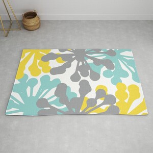 May include: A rectangular rug with a repeating floral pattern in yellow, gray, and teal on a white background. The rug is placed on a gray floor. A gold metal side table and a woven basket are in the background.