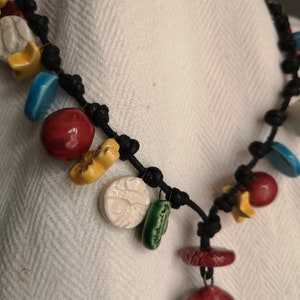 Può includere: Una collana fatta a mano con un cordoncino nero annodato, ornata da ciondoli in ceramica colorati. I ciondoli includono forme rosse, gialle, blu, verdi e bianche, tra cui sfere, stelle e pezzi piatti e rotondi. La collana è esposta su uno sfondo di tessuto bianco.