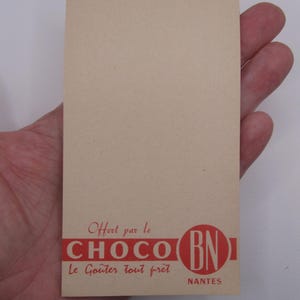May include: A rectangular, light tan card with red text and a red circular logo. The text reads "Offert par le CHOCO Le Goûter tout prêt" and "BN NANTES". The card is held in a hand.