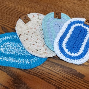 May include: Four crocheted oval rugs with a white border and a brown leather tag with the text "De" on each. The rugs are in shades of blue, white, and beige.