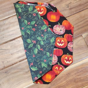 May include: Reversible fabric accessory featuring a Halloween pumpkin pattern on black and a holly berry design on dark green. The accessory is folded, showcasing both sides against a wooden surface.