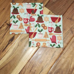 May include: Two square pot holders with a light blue background and a repeating pattern of Christmas baking items. The pattern includes gingerbread men, holly, mugs, and mixing bowls. Each pot holder has a brown loop.