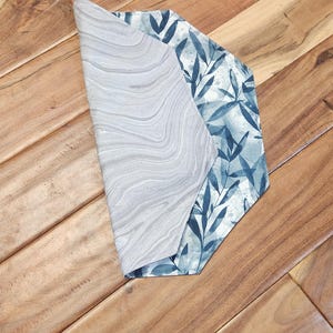 May include: A folded fabric item with two distinct patterns. One side features a gray marble-like design, while the other showcases a blue and white botanical print with leaves. The item is placed on a wooden surface.