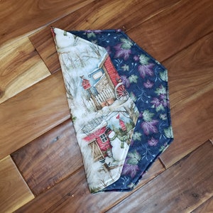 May include: A decorative table runner with a winter scene on one side and a leaf pattern on the other. The winter scene features a snowman, a red building, and a snowy landscape. The leaf pattern is in shades of purple and green.