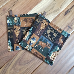 May include: Two brown and green pot holders with a woodland theme. One pot holder features a bear, a cabin, and snowshoes. The other pot holder features a moose, a cabin, and the text "Moose Lake".