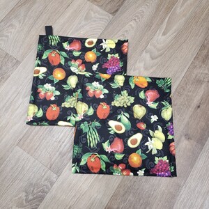 May include: Two black kitchen pot holders with a colorful fruit and vegetable print. The pot holders are made of fabric and have a loop for hanging.