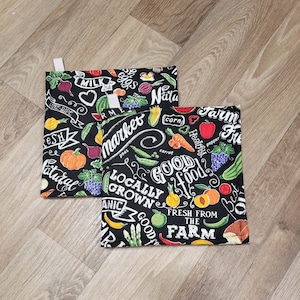 May include: Two black fabric pot holders with a white background and colorful illustrations of fruits and vegetables. The text on the pot holders reads "Farm Fresh", "Locally Grown", "Good Food", and "Market".