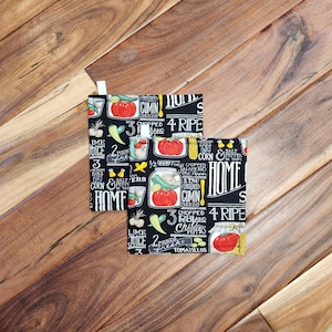 May include: Two black pot holders with a colorful pattern of tomatoes, peppers, and text. The text includes "Cumin Seeds," "Chopped Poblano Chiles," and "Home." The pot holders are on a wooden surface.