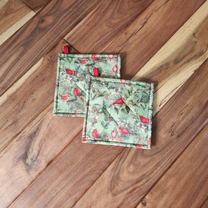 May include: Two green quilted pot holders with a red cardinal and holly pattern. The pot holders have red ribbon loops for hanging.