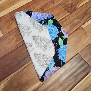 May include: A folded fabric item with three distinct patterns. One side features a white background with gold floral and the text "Merry Christmas". Another side displays blue and purple hydrangeas on a black background. The third side is a solid white.