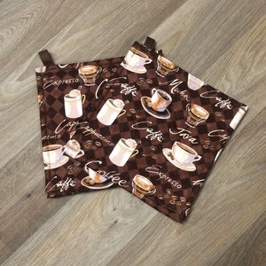 May include: Two brown and white checkered fabric pot holders with a coffee theme. The pot holders feature various coffee drinks, including cappuccino, espresso, java, and mocha, with coffee beans scattered throughout the design.