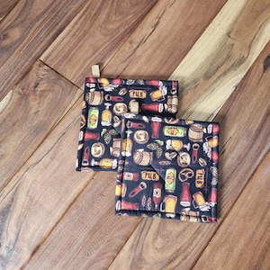 May include: Two black square pot holders with a pattern of beer bottles, mugs, and pub signs. The pot holders have a tan fabric trim.