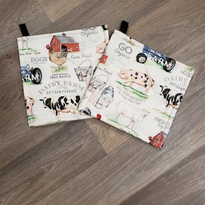 May include: Three white fabric kitchen pot holders with a farm theme. The pot holders feature a red barn, a tractor, cows, pigs, chickens, and the text "Free Range Dairy Farm Artisan Produce", "Go Organic", and "On the Farm".