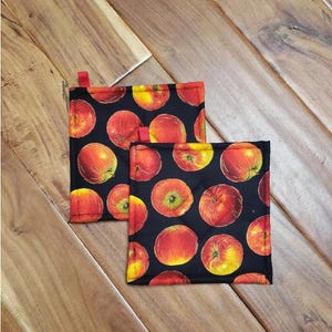 May include: Two black pot holders with a red apple pattern. The pot holders are made of fabric and have a quilted texture.