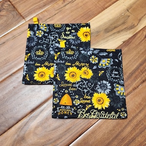 May include: Two black and yellow pot holders with a sunflower and bee design. The pot holders have a repeating pattern of sunflowers, bees, and the words "You are my sunshine", "Sweet as bee", and "Bee grateful". Each pot holder has a yellow loop.
