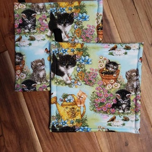 May include: Set of three blue and green kitchen pot holders with a floral pattern and kittens. The kittens are black, white, and orange. The pot holders have a yellow watering can and a bird.