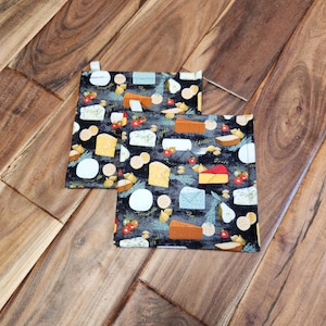 May include: Two black fabric pot holders with a pattern of different types of cheese, grapes, and herbs. The pot holders are on a wood floor.