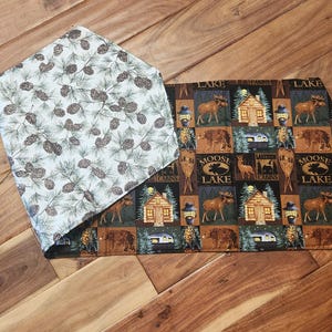 May include: A folded table runner with two distinct patterns. One side features a repeating pattern of pine cones on a light background. The other side has a repeating pattern with moose, cabins, and the words "Moose Lake".